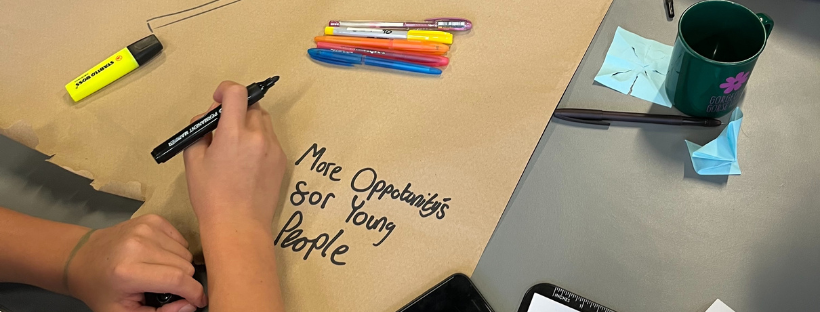 Young people's hands writing on a large piece of brown paper 'More opportunities for young people'