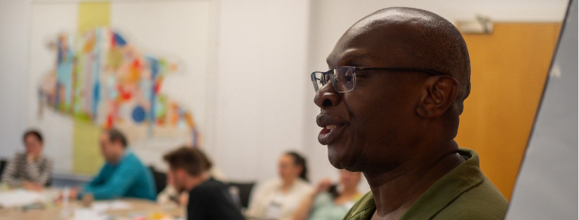 Image of a man facilitating a workshop while a group of people listen in the background.