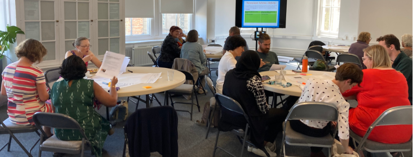 People sitting at different tables in groups, taking part in a workshop. A PowerPoint slide is in the background.