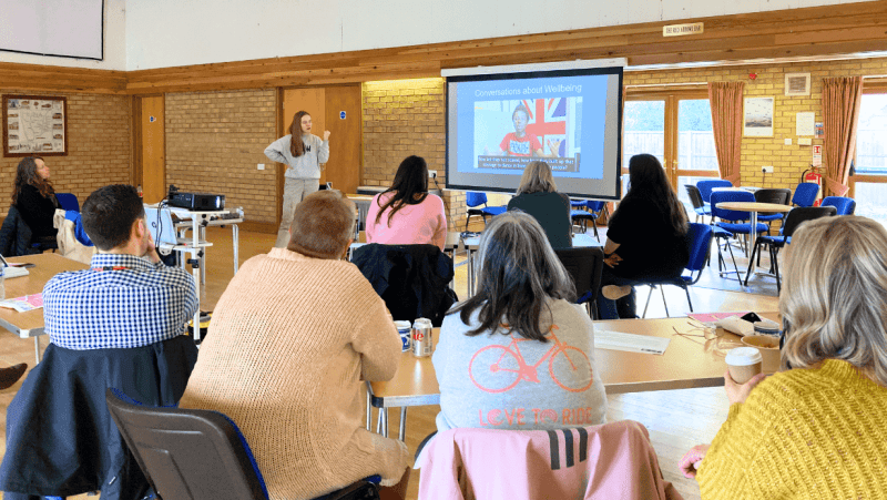 A group of people with their backs to the camera are facing a projection screen with a person with long hair and casual dress standing by it. The training is taking place in a community venue.