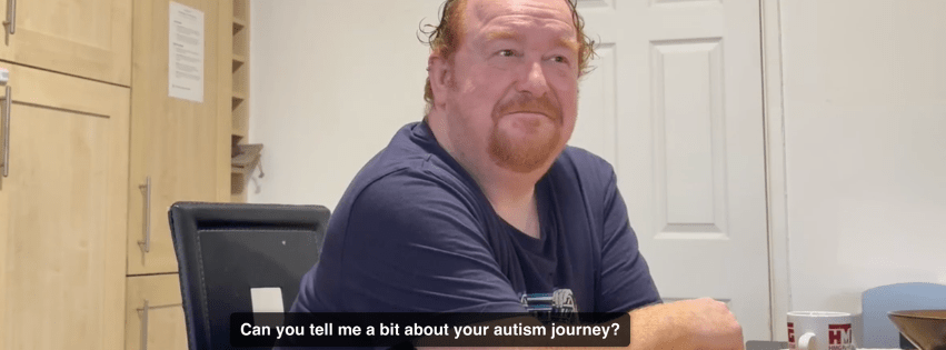 Image is a screen capture from a video interview. The subject is a white man with ginger hair and a goatee beard, wearing a navy tshirt and looking relaxed. A closed caption on the bottom of the image reads "Can you tell me a bit about your autism journey?"