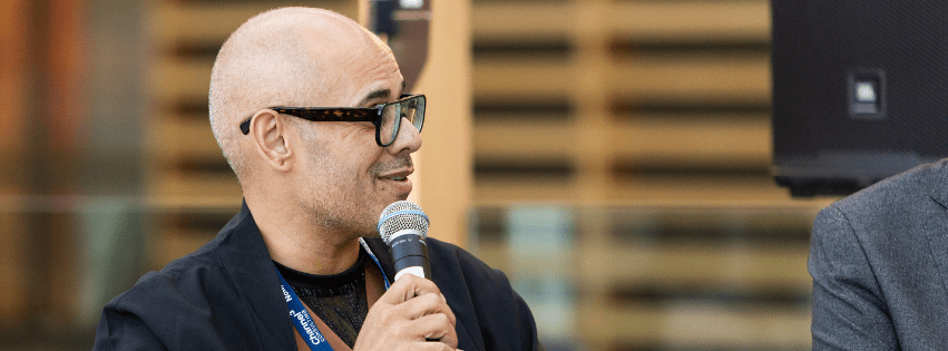 A person with a shaved head and glasses talks into a microhpone